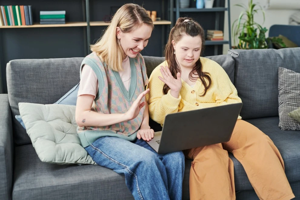 Support worker sitting on couch with disabled woman with down syndrome using laptop